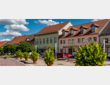 Straßenszene mit historischen, zweistöckigen Häusern mit roten Ziegeldächern und hellen Fassaden, flankiert von kleinen Geschäften und geparkten Autos; im Vordergrund befinden sich große Pflanzkübel mit grünen Pflanzen auf einem gepflasterten Platz unter blauem Himmel mit weißen Wolken.