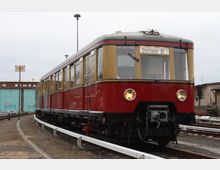 Historischer Zug mit rot-beiger Lackierung auf einem Gleis, beleuchtet mit zwei Frontscheinwerfern. Im Hintergrund stehen ein gemauertes Gebäude mit blauen Toren und ein Schild mit der Aufschrift „1. Halt!“.