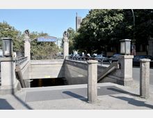Eingang zur U-Bahn-Station Hohenzollernplatz in Berlin, umgeben von massiven Steinpfeilern mit Adlerfiguren und Laternen. Im Hintergrund sind Bäume, parkende Autos und ein modernes Gebäude zu sehen.