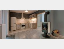 Küche im Landhausstil mit Holzfronten, einer cremefarbenen Kühl-Gefrierkombination, einem Backofen mit Dunstabzugshaube und einem Spülbecken. Im Vordergrund steht ein freistehender Kaminofen mit Holzkorb daneben, auf hellem Laminatboden.