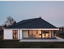 Modernes Einfamilienhaus mit flachem Satteldach und großen Glasfronten, die einen Blick in den beleuchteten Wohn- und Essbereich ermöglichen; umgeben von einer Grasfläche und angrenzenden Wohnhäusern in der Ferne.