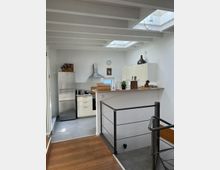 Moderne Küche mit weißen Schränken, Edelstahlgeräten und einer Arbeitsplatte aus Holz. Der Raum hat sichtbare Holzbalken an der Decke, große Dachfenster, die viel Licht einlassen, und einen angrenzenden Treppenaufgang mit Metallgeländer.