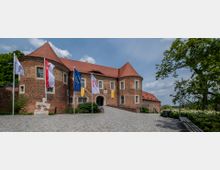 Historisches Backsteingebäude mit roten Ziegeldächern und runden Türmen, umgeben von einem gepflasterten Vorplatz mit wehenden Flaggen. Im Vordergrund stehen Bänke und Bäume, während der Eingang durch ein großes Tor in der Mitte des Gebäudes hervorgehoben wird.