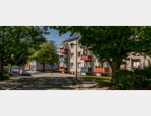 Wohnstraße mit einem Mehrfamilienhaus auf der rechten Seite, dessen Balkone mit roten und blauen Paneelen dekoriert sind, umgeben von großen Bäumen und Grünflächen. Im Vordergrund eine gepflasterte Straße mit parkenden Autos und Laternen.