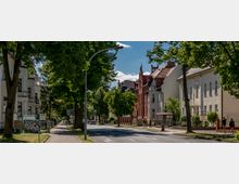 Straßenansicht eines Wohnviertels mit Bäumen, Bürgersteigen und historisch anmutenden Gebäuden, darunter ein rotes Backsteinhaus mit dekorativen Giebeln. Auf der rechten Seite befindet sich eine Bushaltestelle, umgeben von wenig Verkehr und Fußgängern.