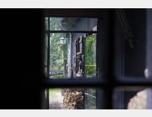 Blick durch ein Fenster auf einen Außenbereich mit einer dunklen Hausfassade, Holzstapeln, geschnitzten Figuren an der Wand und einer Laterne. Im Hintergrund sind Bäume und grüne Vegetation sichtbar.
