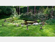 Ein Garten mit einer kleinen Teichlandschaft, die mit Seerosen bedeckt ist, umgeben von dichter Vegetation und Bäumen. Im Vordergrund stehen mehrere künstlerische Skulpturen auf Metallgestellen auf einer grasbewachsenen Fläche.