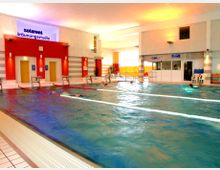 Hallenbad mit mehreren Schwimmbahnen, umgeben von einer hellen Innenraumgestaltung mit rot-weiß gestreiften Wänden und einem Bereich mit Türen sowie Schildern. Im Hintergrund befinden sich ein Fensterbereich, ein Tresen sowie Stühle und bunte Schwimmutensilien.