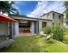 Moderner Wohnhaus-Anbau mit großer, aufgeklappter Glasfront zur Terrasse, die den Blick in eine offene Küche mit Esstisch freigibt. Im Vordergrund liegt ein Rasen mit zwei Lounge-Stühlen und einem Sonnenschirm, daneben ein dicht bepflanztes Gartenbeet.
