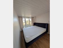 Schmales Schlafzimmer mit hellen Holzpaneelen an Wänden und Decke, Holzboden und einem großen Bett mit dunklem Polsterkopfteil. Am Ende des Raums befindet sich eine bodentiefe Glasschiebetür mit Vorhängen und Blick ins Grüne.