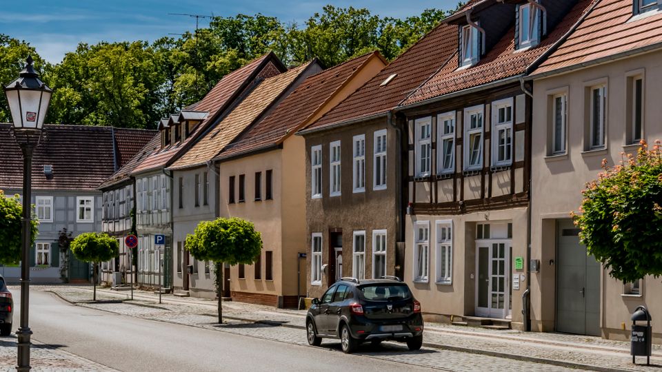 Straßenzug in einer Kleinstadt mit Kopfsteinpflaster, parkenden Autos und kugelförmig beschnittenen Straßenbäumen. Entlang der Straße stehen eng gereihte, zweigeschossige Wohnhäuser mit Putzfassaden und teils Fachwerkdetails unter roten Satteldächern.