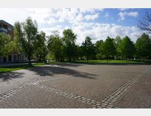 Ein gepflasterter Platz mit quadratischen Mustern, umgeben von Bäumen und Grünflächen. Im Hintergrund befindet sich ein modernes Gebäude mit Balkonen sowie ein Gehweg neben einer parkähnlichen Landschaft.