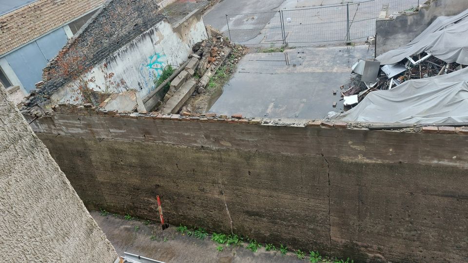 Eingezäuntes, verlassenes Gelände mit zerfallenden Mauerresten, Trümmern und einer Betonfläche, teilweise bedeckt mit Pfützen. Im Hintergrund liegen Schutthaufen unter grauen Planen.