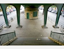 Treppengang unter einer grünen, stählernen Hochbahnbrücke mit Pfeilern und strukturiertem Boden aus Kopfsteinpflaster. Im Zentrum steht ein kleines, rundes Gebäude mit einem grünen Eingang, dahinter führt eine Straße mit parkenden und fahrenden Autos entlang.