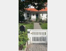 Weißes Einfamilienhaus mit rotem Ziegeldach, grün gestrichener Haustür und symmetrischen Fenstern. Ein gepflasterter Weg führt durch einen Vorgarten mit Rasen und Pflanzen zur Eingangstür, umgeben von einem niedrigen weißen Zaun mit Gartentor.