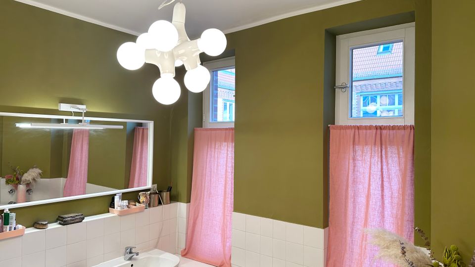 Badezimmer mit olivgrünen Wänden, weißer Fliesenverkleidung und zwei Waschbecken unter einem breiten Spiegel mit Leuchtleiste. Zwei hohe Fenster mit rosa Vorhängen und eine moderne Deckenleuchte mit mehreren Kugellampen prägen den Raum.