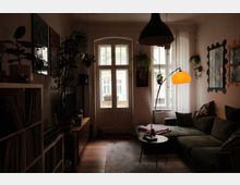 Gemütliches Wohnzimmer mit hohen Fenstern und einer Glastür zum Balkon, davor ein Sofa mit Couchtisch und Stehlampe. Pflanzen hängen an der Decke und an den Wänden; links stehen Regale und ein Lowboard, rechts Wandspiegel und Bilder.