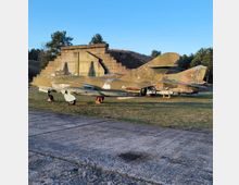 Zwei veraltete Militärflugzeuge in Tarnfarben stehen auf einer Wiese neben einem betonartigen, abgestuften Bauwerk. Im Hintergrund sind Bäume und ein klarer Himmel sichtbar.