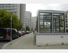 Straßenszene mit hohen Wohngebäuden aus Beton und Glas, links geparkte Autos entlang eines Gehwegs mit Bäumen, und rechts eine gläserne, moderne Struktur mit schwarzen Metallrahmen. Im Hintergrund befinden sich weitere Hochhäuser.