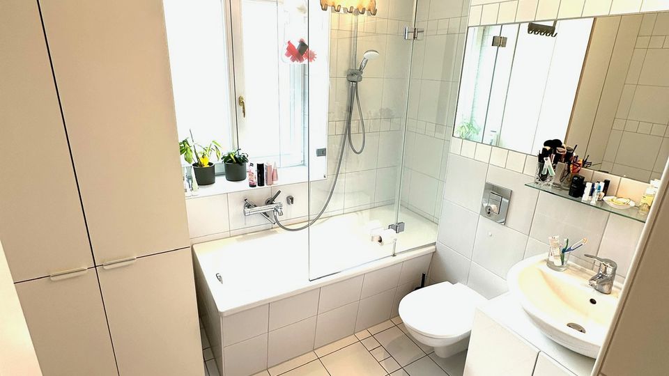 Helles, weiß gefliestes Badezimmer mit Badewanne und Glas-Duschabtrennung unter einem großen Fenster mit Rundbogen. Rechts befinden sich ein wandhängendes WC, ein Waschtisch mit Spiegel sowie ein hoher Schrank; der Boden ist mit großen quadratischen Fliesen ausgelegt.