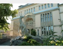 Historisches Gebäude mit der Aufschrift "Aquarium" über dem Eingangsportal, mit neoklassizistischer Architektur und verzierten Fenstern. Im Vordergrund steht eine große steinerne Echsenstatue auf einem Felsen, umgeben von Bäumen und Vegetation.