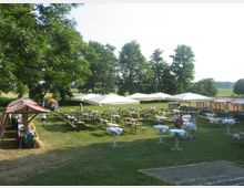 Eine Freiluftveranstaltung auf einer Wiese mit langen Bierbänken und Tischen, teils unter weißen Sonnenschirmen. Im Hintergrund stehen Bäume, und rechts sowie links sind Marktstände mit rot-weiß gestreiften Dächern zu sehen.