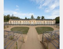 Historisches Gebäude im barocken Stil, umgeben von einem symmetrisch angelegten Garten mit Rasenflächen und Sandwegen, sowie Pergolen aus Holz. Das Gebäude hat eine helle Fassade, große Fenster und einen zentralen Uhrturm mit grüner Kuppel.