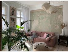 Ein gemütliches Wohnzimmer mit hellen Holzfußböden, einer rosa Stoffcouch und einer alten, teils abblätternden Wand in Pastellgrün. Große Fenster mit weißen Vorhängen, Zimmerpflanzen, dekorative botanische Poster und eine weiße Lampe mit Papierenschirm ergänzen die entspannte Atmosphäre.