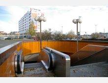 Eingang zu einer U-Bahn-Station mit orangefarbenen Kacheln und Rolltreppen, umgeben von einer urbanen Umgebung mit Straßenlaternen und einem mehrstöckigen Hotelgebäude mit der Aufschrift „Hotel ibis“ im Hintergrund.