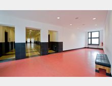 Geräumiger, heller Innenraum mit einem roten Bodenbelag, weißen Wänden und schwarzen Fliesen an der unteren Wandhälfte. Links führen offene Durchgänge in einen langen Flur mit schwarzem und beige kariertem Boden, während rechts ein großes Fenster natürliches Licht hereinlässt; eine schwarze gepolsterte Sitzbank steht am Rand des Raumes.