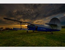 Ein kleines, blau-weißes Propellerflugzeug steht auf einer grasbewachsenen Fläche bei Sonnenuntergang, mit dramatisch bewölktem Himmel im Hintergrund.