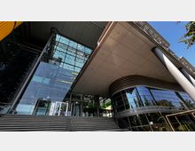 Moderne Gebäudefassade mit einer Kombination aus großflächigen Glasfronten und glatten, überstehenden Dachkonstruktionen. Im Eingangsbereich sind eine Treppe, Glasdrehtüren und markante Metallstützpfeiler erkennbar.