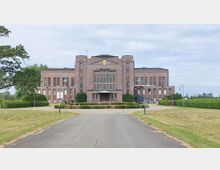 Klinkergebäude im Stil des frühen 20. Jahrhunderts mit symmetrischer Fassade, hohen Fenstern und einem überdachten Eingangsbereich, umgeben von einer gepflegten Grünanlage mit Bäumen, Wegen und Rasenflächen. Im Vordergrund führt ein asphaltierter Weg zum Gebäude.