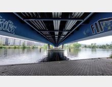 Unteransicht einer modernen Brücke mit blauer Stahlkonstruktion, die über einen Fluss führt. An den Brückenpfeilern und der Konstruktion sind Graffiti sichtbar, im Hintergrund befinden sich Wohngebäude und grüne Uferbereiche.