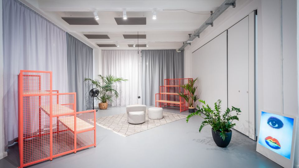 Heller, moderner Studio- oder Galerieraum mit grauem Boden, weißen Wänden und Akustikpaneelen sowie Schienenleuchten an der Decke. Im Raum stehen orangefarbene Gitterregale, mehrere Topfpflanzen und zwei runde Sitzhocker auf einem Teppich; an der Wand lehnt ein beleuchtetes Kunstbild.