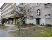 Mehrstöckiges Wohngebäude mit grauer Putzfassade, schmalen Fenstern und einer überdachten Durchfahrt. Im Vordergrund befindet sich eine Grünfläche mit kahlen Bäumen und Gehwegen, daneben steht ein Fahrrad an der Eingangstür des Hauses.