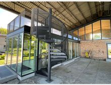 Innenansicht eines modernen Gebäudes mit industriellem Stil, das große Glasflächen und eine freistehende Wendeltreppe mit Metallgitter umfasst. Die Umgebung beinhaltet unverputzte Ziegelwände, ein sichtbares Stahldach und Blick ins Grüne durch die Fenster.