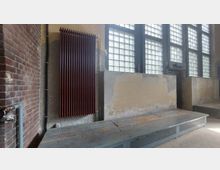 Industrieähnlicher Innenraum mit einem roten Heizkörper an einer beigen Wand, daneben eine braune Backsteinwand. Im Hintergrund befinden sich hohe Fenster mit Glasbausteinen, die viel Licht hereinlassen, sowie eine erhöhte Metallplattform im Vordergrund.