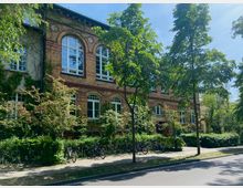 Backsteingebäude im historischen Stil mit großen, bogenförmigen Fenstern, umgeben von Bäumen und Sträuchern. Auf dem Gehweg vor dem Gebäude stehen Fahrräder, und die Szenerie ist von Tageslicht beleuchtet.