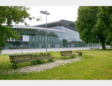 Ein modernes Gebäude mit weitläufiger Glasfassade und geschwungenem Dach liegt im Hintergrund, während im Vordergrund mehrere Holzbänke auf einer Rasenfläche stehen, umgeben von Bäumen und gepflasterten Wegen.