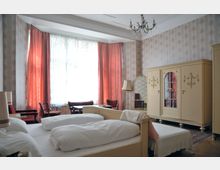 Ein klassisch eingerichtetes Schlafzimmer mit zwei Einzelbetten, einem cremefarbenen Kleiderschrank, einer Stehlampe und einem Wandspiegel. Große Fenster mit Vorhängen und floraler Tapete verleihen dem Raum einen nostalgischen Charakter.