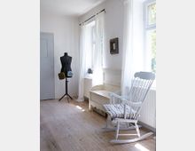 Heller Raum mit Holzboden, einer Schneiderpuppe mit Taschen, einem weißen Schaukelstuhl mit Kissen und einer Sitzbank unter hohen Fenstern mit weißen, halbtransparenten Vorhängen.