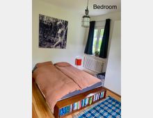 Schlafzimmer mit einem Holzbett, dessen Fußende als Regal für Bücher genutzt wird. An der Wand hängt ein großformatiges Schwarz-Weiß-Bild, rechts daneben sind ein Fenster mit dunklen Vorhängen und eine weiße Heizung zu sehen.