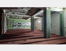 Innenraum eines verlassenen Gebäudes mit terrassenförmig ansteigendem Boden, mehreren grünen Stützpfeilern und einer stark abblätternden Decke. Wände mit Graffiti und verwitterter Farbe, seitlich eine Reihe hoher Fenster und einfache Türen.
