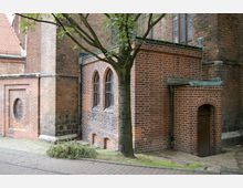 Backsteinfassade eines historischen Gebäudes mit gotischen Elementen, wie spitzbogigen Fenstern mit Metallgittern. Im Vordergrund steht ein Baum, und rechts unten ist eine kleine Holztür in einer gerundeten Nische sichtbar.