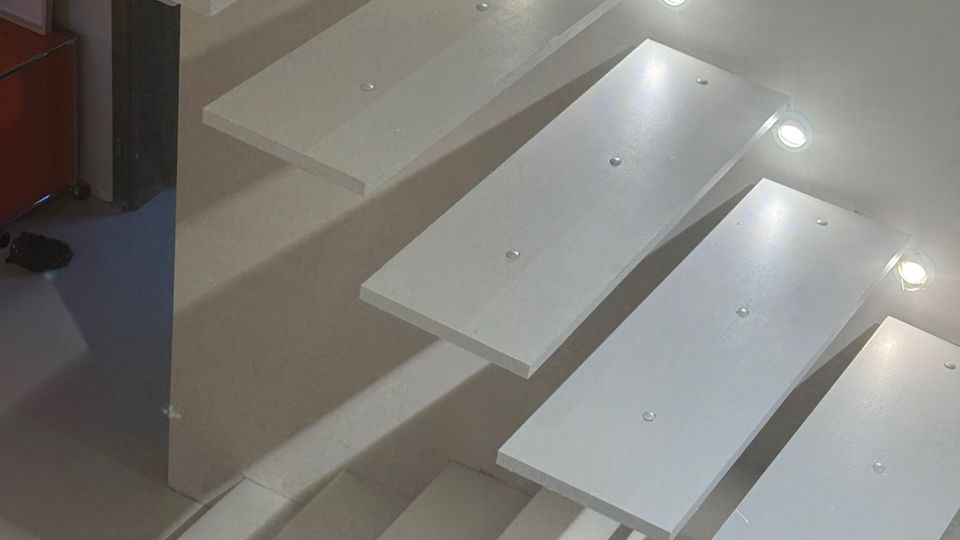 Interior view of a modern staircase with white floating rectangular steps attached to a wall, lit by small round recessed lights along the side. The steps cast strong linear shadows onto the lower landing and wall.