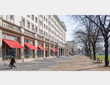 Straßenansicht eines mehrstöckigen Gebäudes mit heller Fassade und großen Fenstern, vor denen rote Markisen hängen. Rechts daneben verläuft ein breiter Gehweg mit Bäumen und einer angrenzenden Grünfläche, mit einzelnen Bänken und wenigen Passanten.