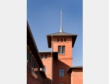 Backstein-Gebäude mit Turm und spitzem Dach sowie großen Fenstern, umgeben von klarem, blauem Himmel; auf der linken Seite ist ein Teil eines angrenzenden Gebäudes mit Glasfenstern sichtbar.