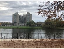 Großes modernes Betongebäude mit mehreren Etagen steht in der Mitte des Bildes, umgeben von Bäumen und einer Wiese. Im Vordergrund befindet sich ein ruhiger Fluss mit einem Metallgeländer und herbstlichem Laub am Ufer.