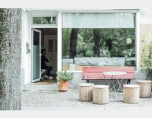 Straßenszene vor einem kleinen Café mit großer Schaufensterscheibe, einer roten Holzbank, einem runden Metalltisch und mehreren zylinderförmigen Hockern aus Holz. Ein geöffneter Eingang zeigt eine Person im Inneren; davor steht ein Pflanztopf mit Kräutern.